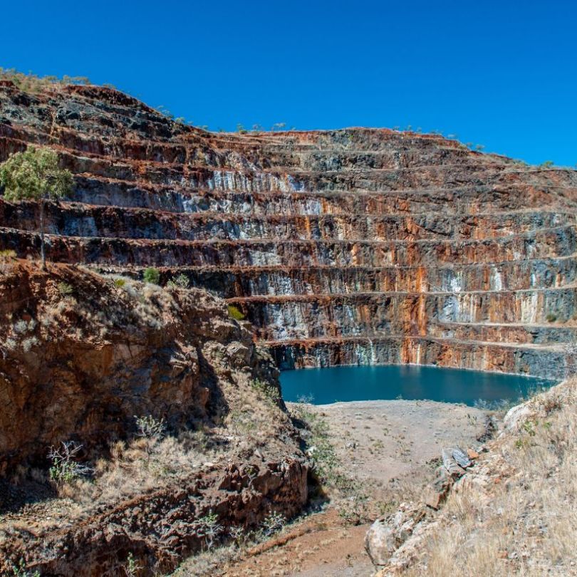An open pit mine with terraced rock layers in varied colors and a small body of water at the bottom, surrounded by dry, rocky terrain.