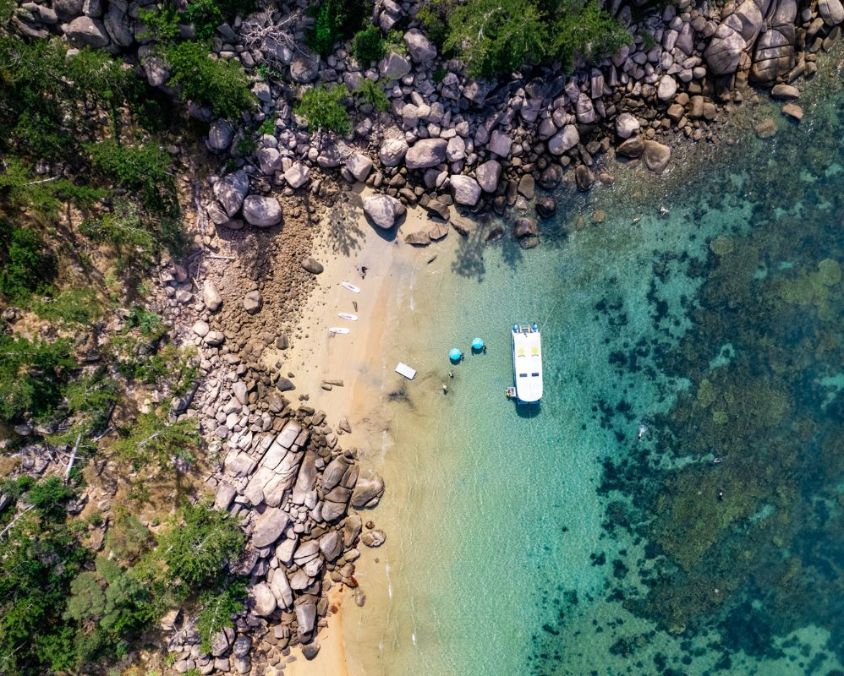 Aerial view of a small sandy beach with turquoise water, a white boat anchored offshore, and kayaks and paddleboards lined up on the shore. Rocky boulders and lush green trees surround the beach, with coral visible beneath the clear water.