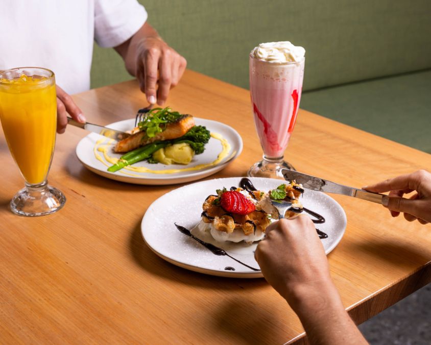 Two plates of food on a wooden table, with two beverages. One with a chocolate-drizzled waffle with a strawberry milkshake, and the other is grilled fish over vegetables with juice