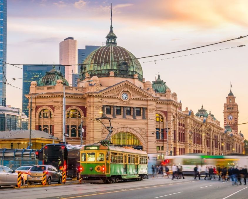Flinders Street Station in Melbourne with trams, vehicles, and pedestrians at the intersection during sunrise or sunset.